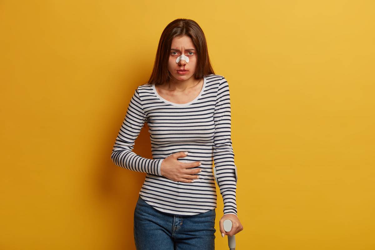 Sad young woman suffers from terrible pain in stomach, injured after road accident or falling down on asphalt from vehicle, poses with crutch, learns to walk after trauma. Rehabilition period